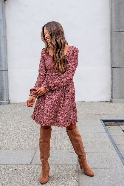 Woman in the Wild Rust Print Smocked Mini Dress - FINAL SALE smiles in tan knee-high boots against a white wall and gray tiled pavement.