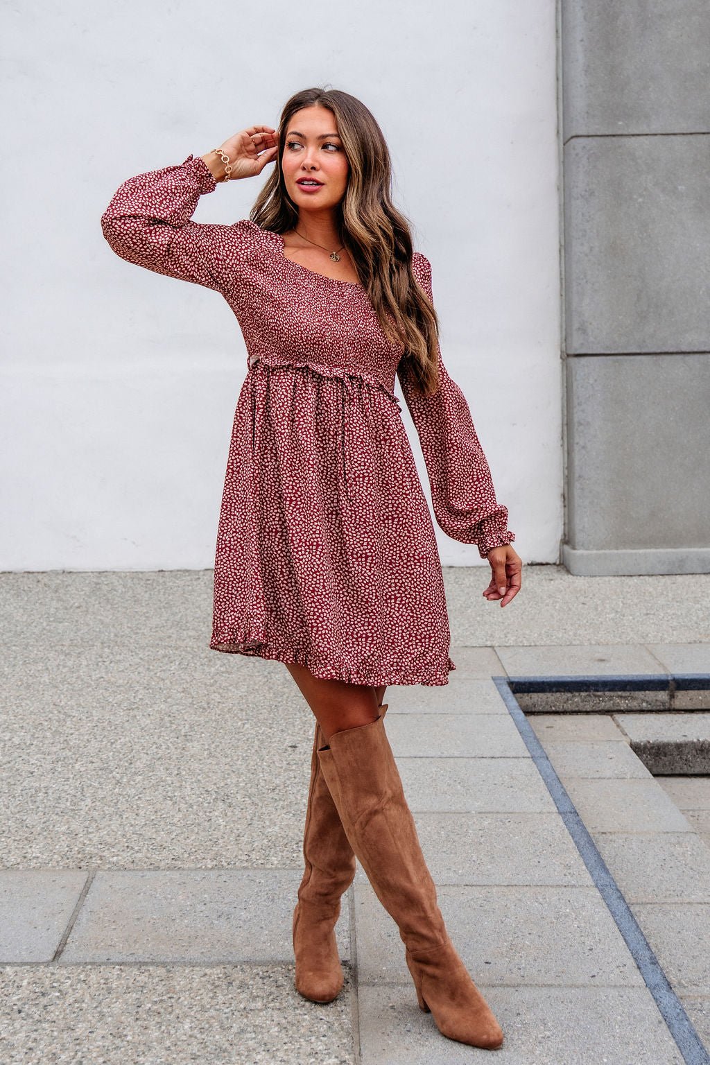Woman in a Magnolia exclusive Wild Rust Print Smocked Mini Dress - FINAL SALE stands outdoors in tan knee-high boots, looking to the side.