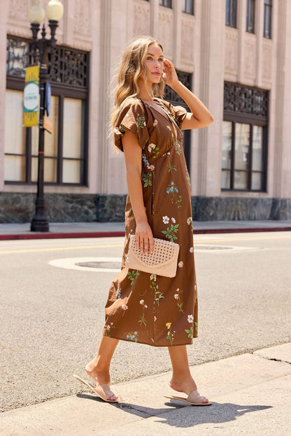 Woman in the Wildflower Floral Midi Dress - Brown walks on a city sidewalk, wearing sandals, with a woven tan purse and touching her hair.