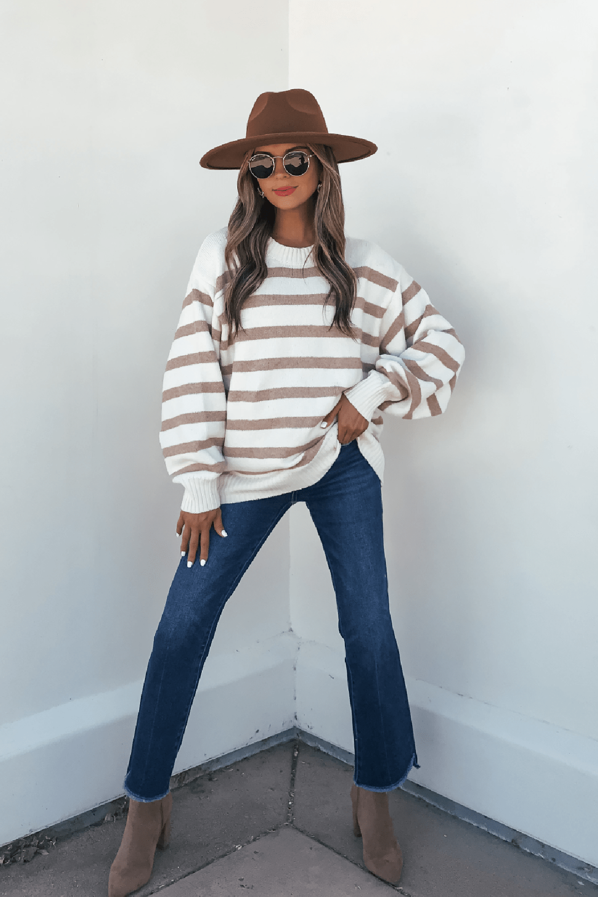 Woman in a brown hat, sunglasses, Willow Cream Stripe Ribbed Sweater, blue jeans, and tan boots stands by a white corner wall.