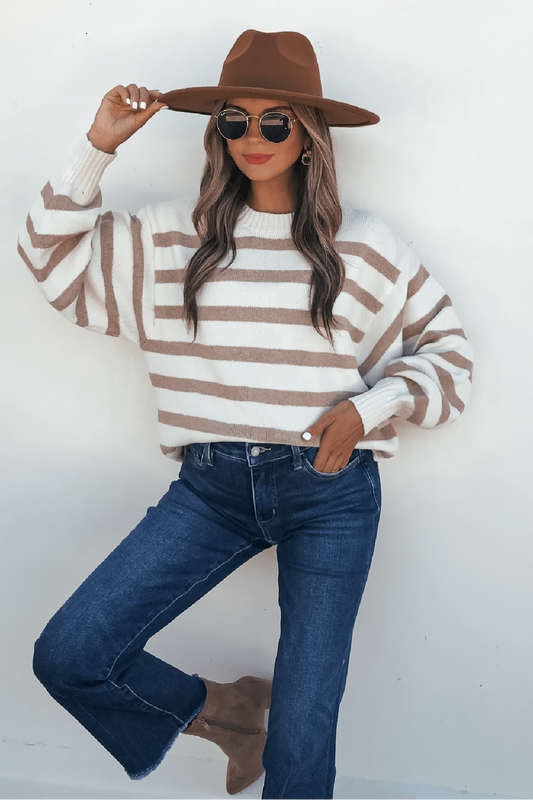 Woman wearing a brown hat, sunglasses, Willow Cream Stripe Ribbed Sweater, blue jeans, and ankle boots poses by a white wall.