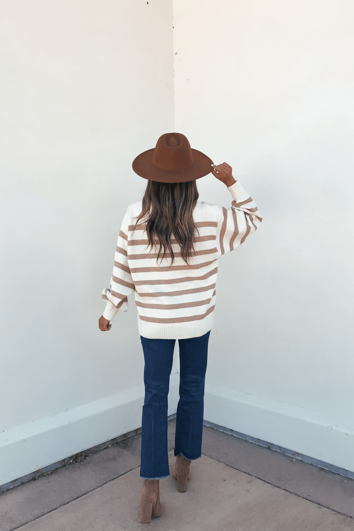 A woman in a brown hat, Willow Cream Stripe Ribbed Sweater, blue jeans, and heeled boots stands facing a white wall from behind.