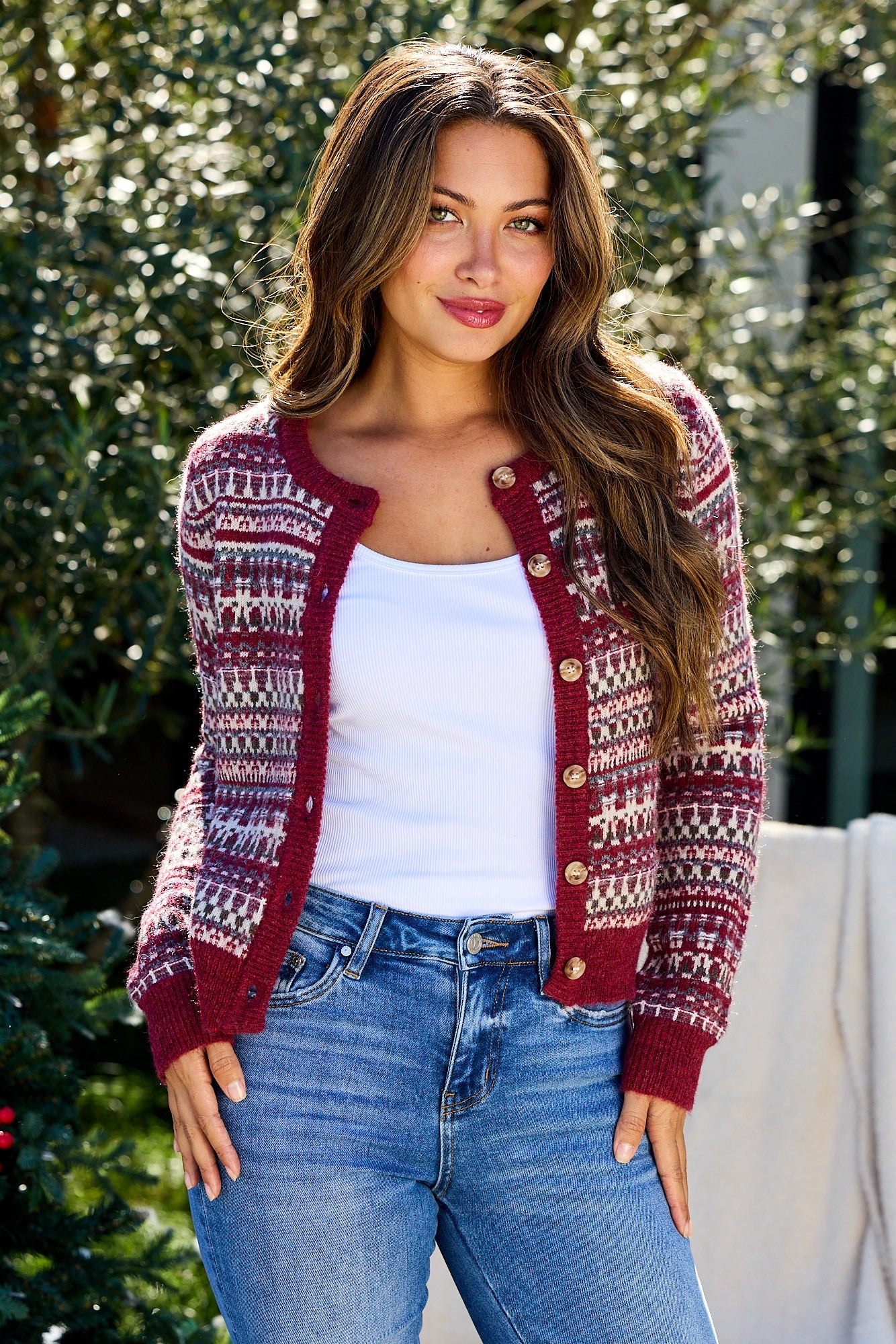 Woman with long brown hair wears a Wine Fair Isle Button Down Cardigan, white tank top, and blue jeans outdoors, smiling.