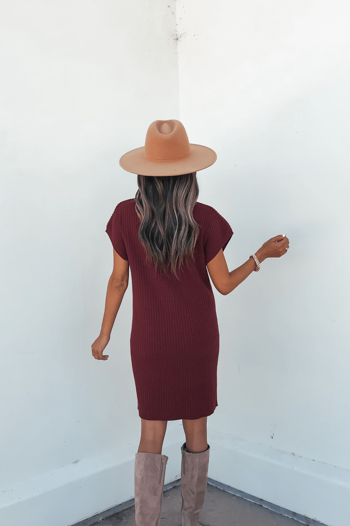 A woman wearing the Wine Mock Neck Mini Sweater Dress stands with her back to the camera, facing a white wall.