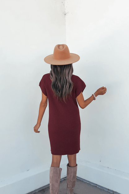 A woman wearing the Wine Mock Neck Mini Sweater Dress stands with her back to the camera, facing a white wall.