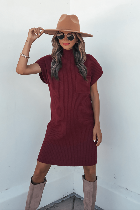 Woman in a Wine Mock Neck Mini Sweater Dress poses against a white wall, accessorized with a hat, sunglasses, and beige boots.