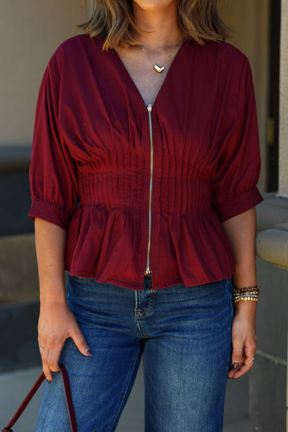 Woman in Wine Ruched Zip Up Flare Top - FINAL SALE, V-neck, with blue jeans and purse, standing outdoors.