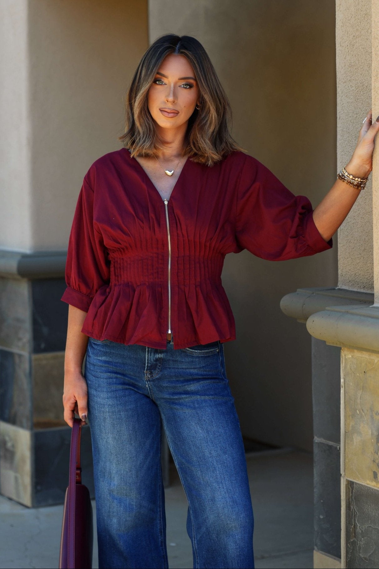 Woman with shoulder-length hair wears the Wine Ruched Zip Up Flare Top - FINAL SALE and blue jeans, standing by a building wall.
