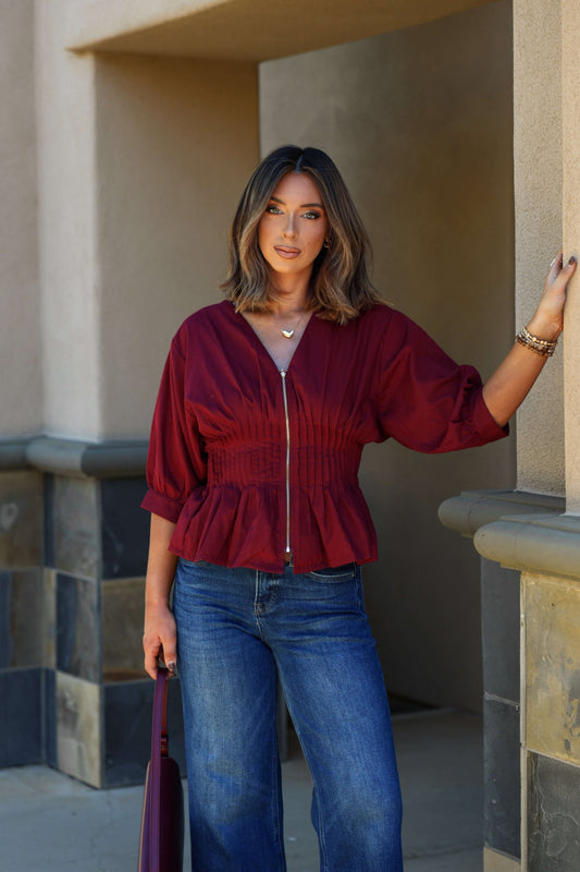 Woman with shoulder-length brown hair wears a Wine Ruched Zip Up Flare Top and blue jeans, holding a matching bag outdoors.