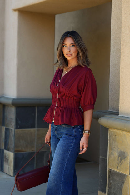 Woman with shoulder-length brown hair in the Wine Ruched Zip Up Flare Top - FINAL SALE, holding a maroon handbag outdoors.