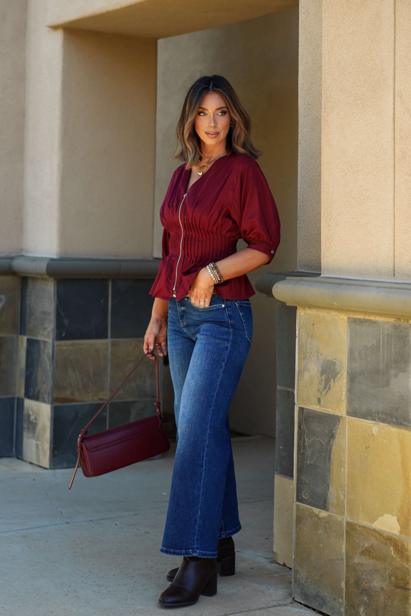 A woman in the Wine Ruched Zip Up Flare Top and blue jeans stands outdoors, smiling with a matching maroon purse and heeled boots.
