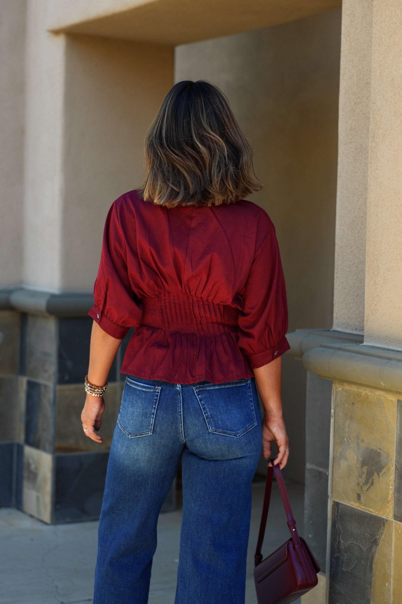 Person in a Wine Ruched Zip Up Flare Top - FINAL SALE and blue jeans, seen from behind, holding a maroon handbag outdoors.