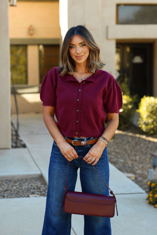 A woman wears a Wine Short Sleeve Button Down Sweater and blue jeans, holding a matching purse and smiling outside.