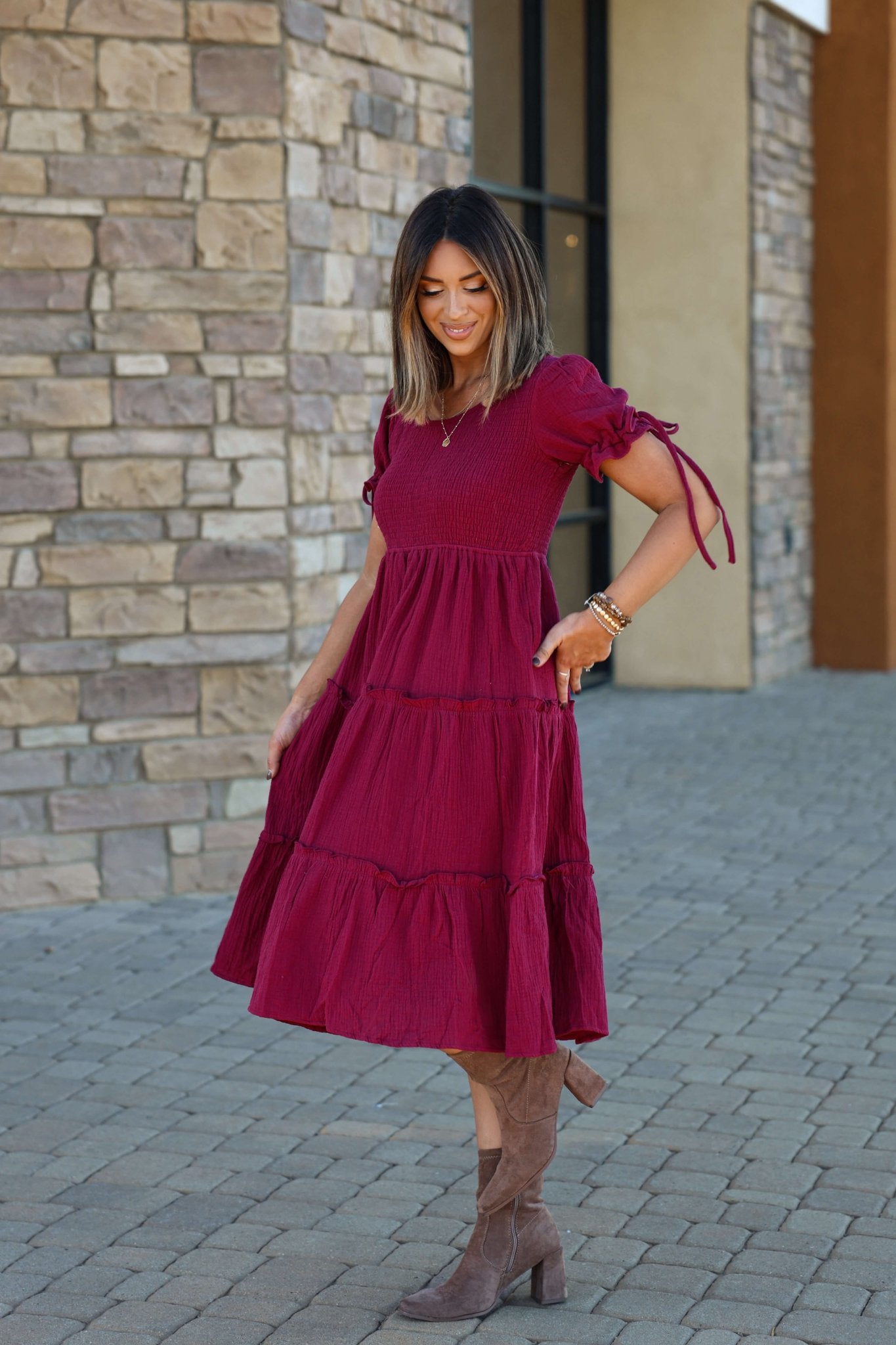 A woman in the Wine Smock Tiered Midi Dress and brown boots smiles, holding the ruffle-tiered skirt on a stone walkway.