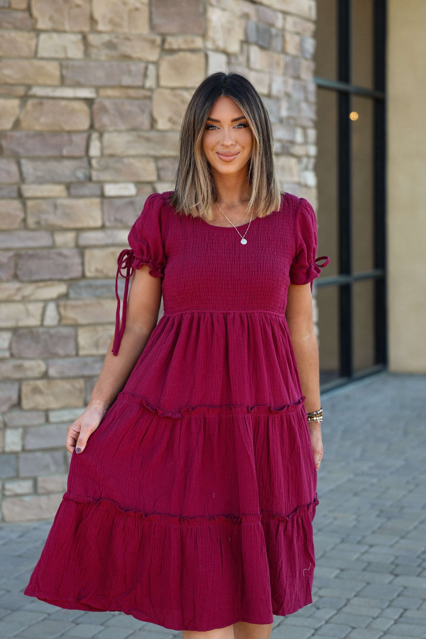 A woman smiles outside on a stone walkway, wearing the Wine Smock Tiered Midi Dress in front of a stone wall.