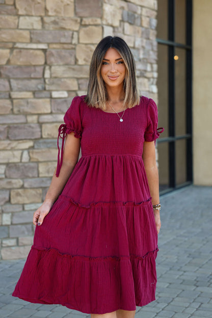 A woman smiles outside on a stone walkway, wearing the Wine Smock Tiered Midi Dress in front of a stone wall.