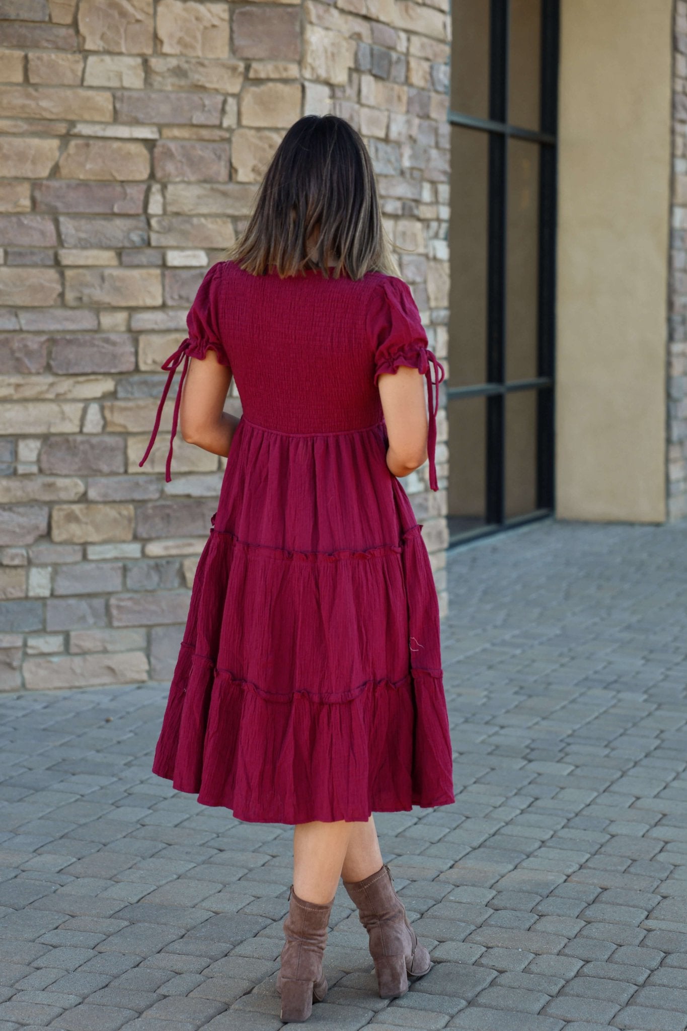 Wearing a Wine Smock Tiered Midi Dress and brown boots, a woman stands on a stone path facing a brick wall, back to camera.