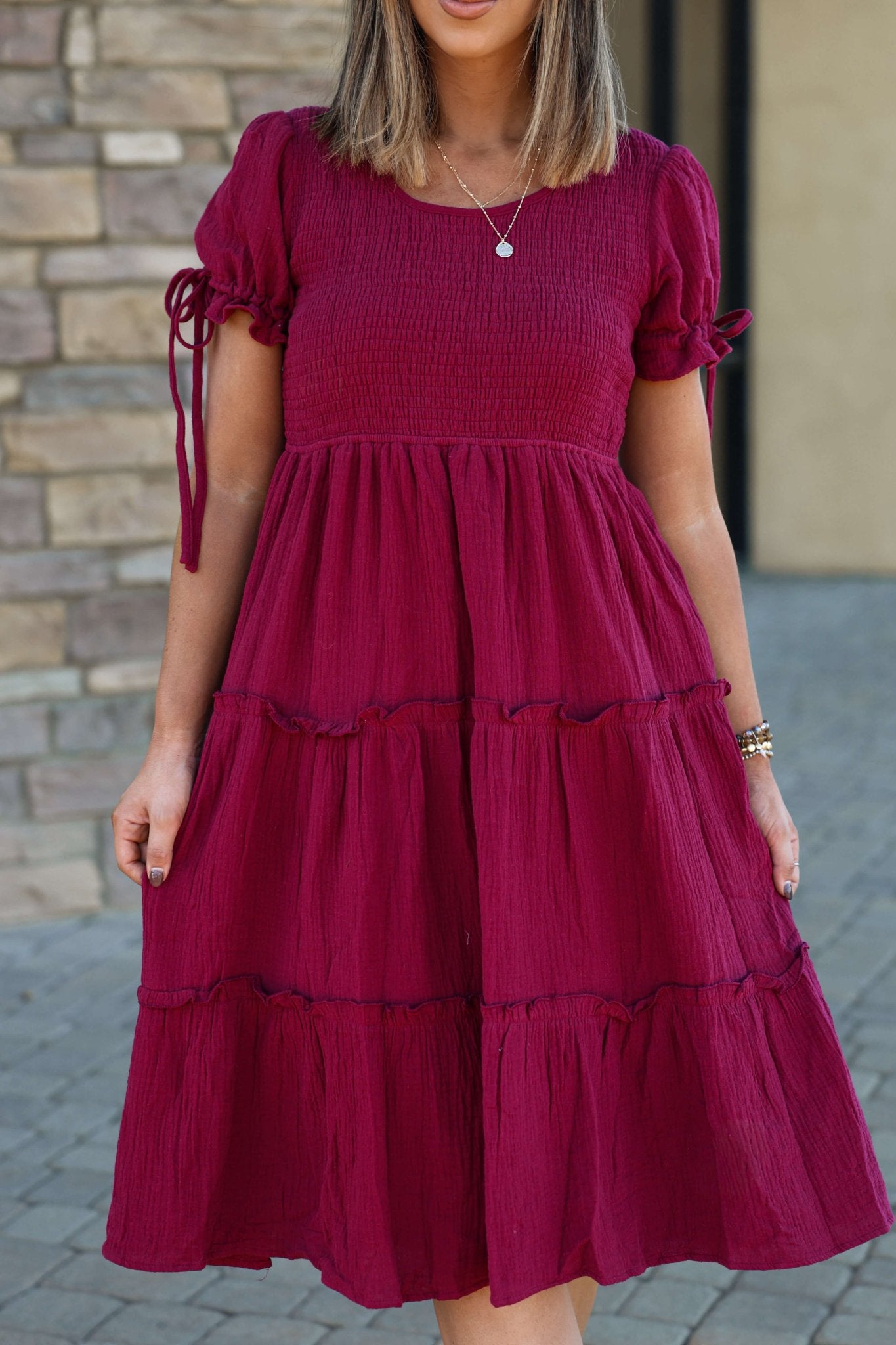 A woman in a Wine Smock Tiered Midi Dress with a ruffled skirt stands outside by a stone wall.