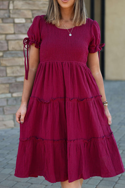 A woman in a Wine Smock Tiered Midi Dress with a ruffled skirt stands outside by a stone wall.
