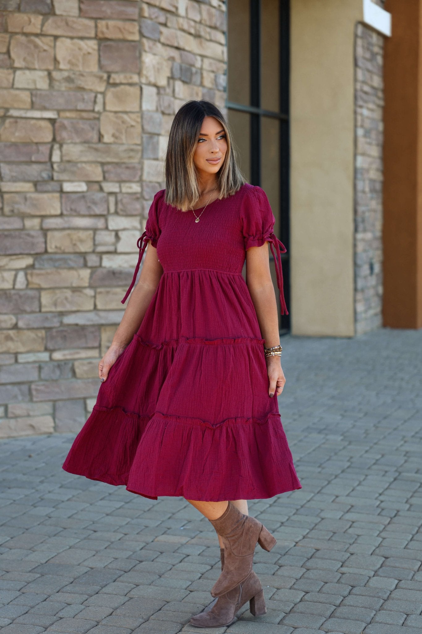 A woman in the Wine Smock Tiered Midi Dress and brown ankle boots stands on a stone walkway before a stone wall.