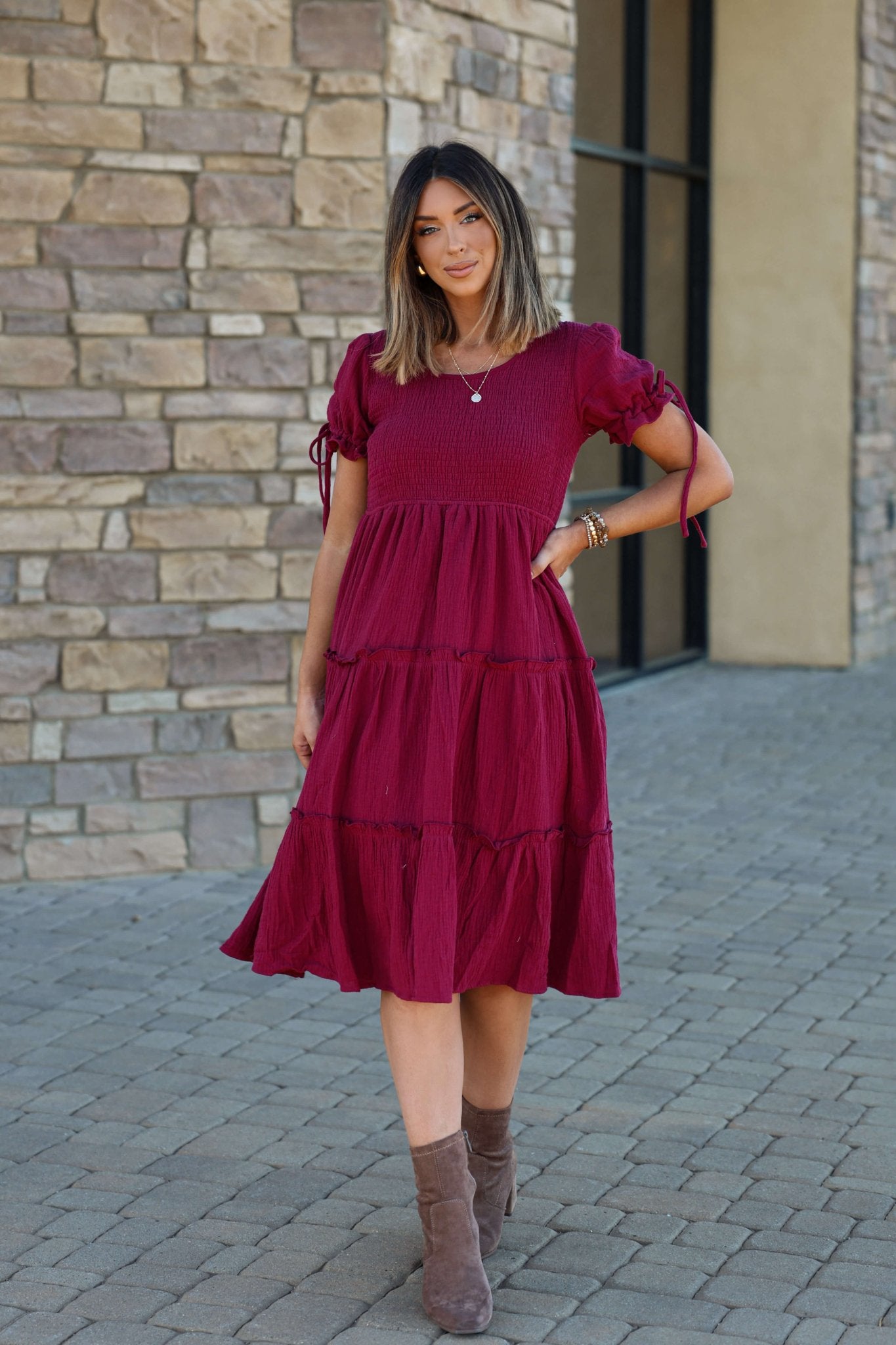 A woman in the Wine Smock Tiered Midi Dress stands on a stone walkway by a brick wall.