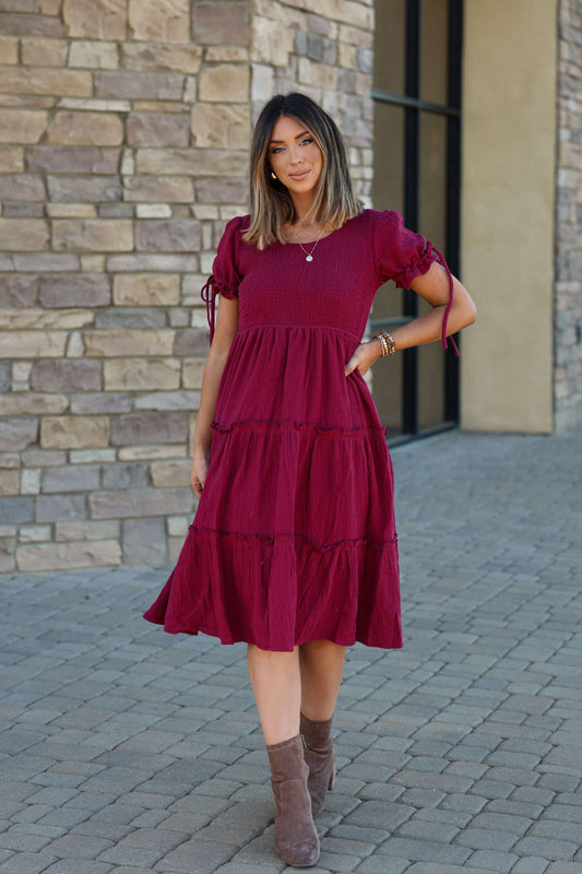 A woman in the Wine Smock Tiered Midi Dress stands on a stone walkway by a brick wall.