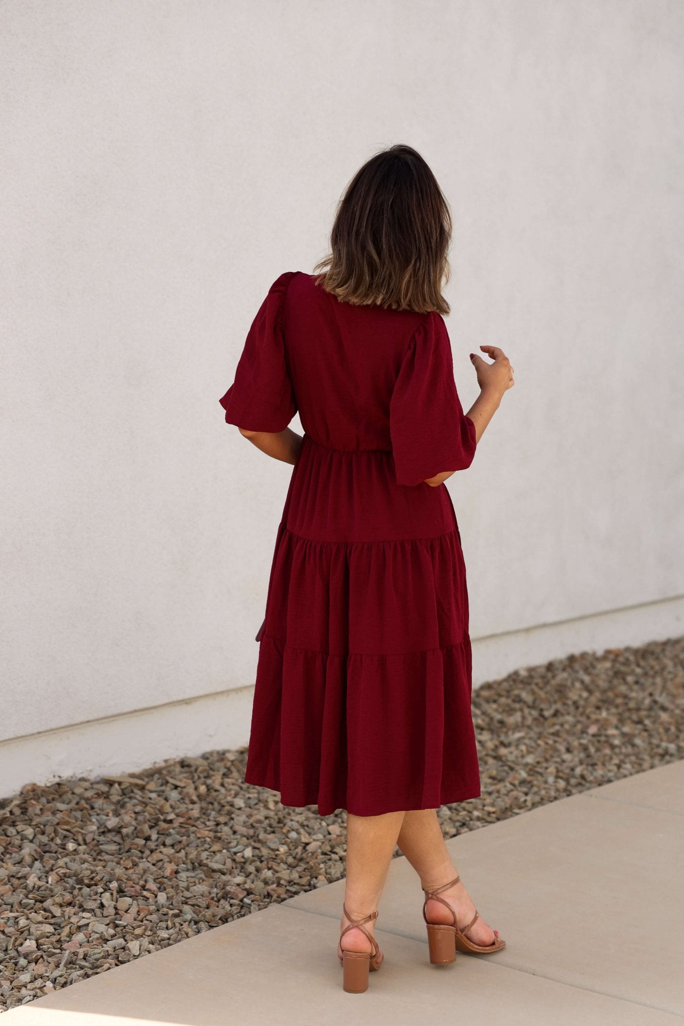 Woman in the Wine Tiered Midi Wrap Dress stands on the sidewalk, its maroon hue and short sleeves visible from behind.