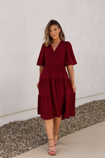 A woman in the Wine Tiered Midi Wrap Dress stands on a stone path beside a light-colored wall.