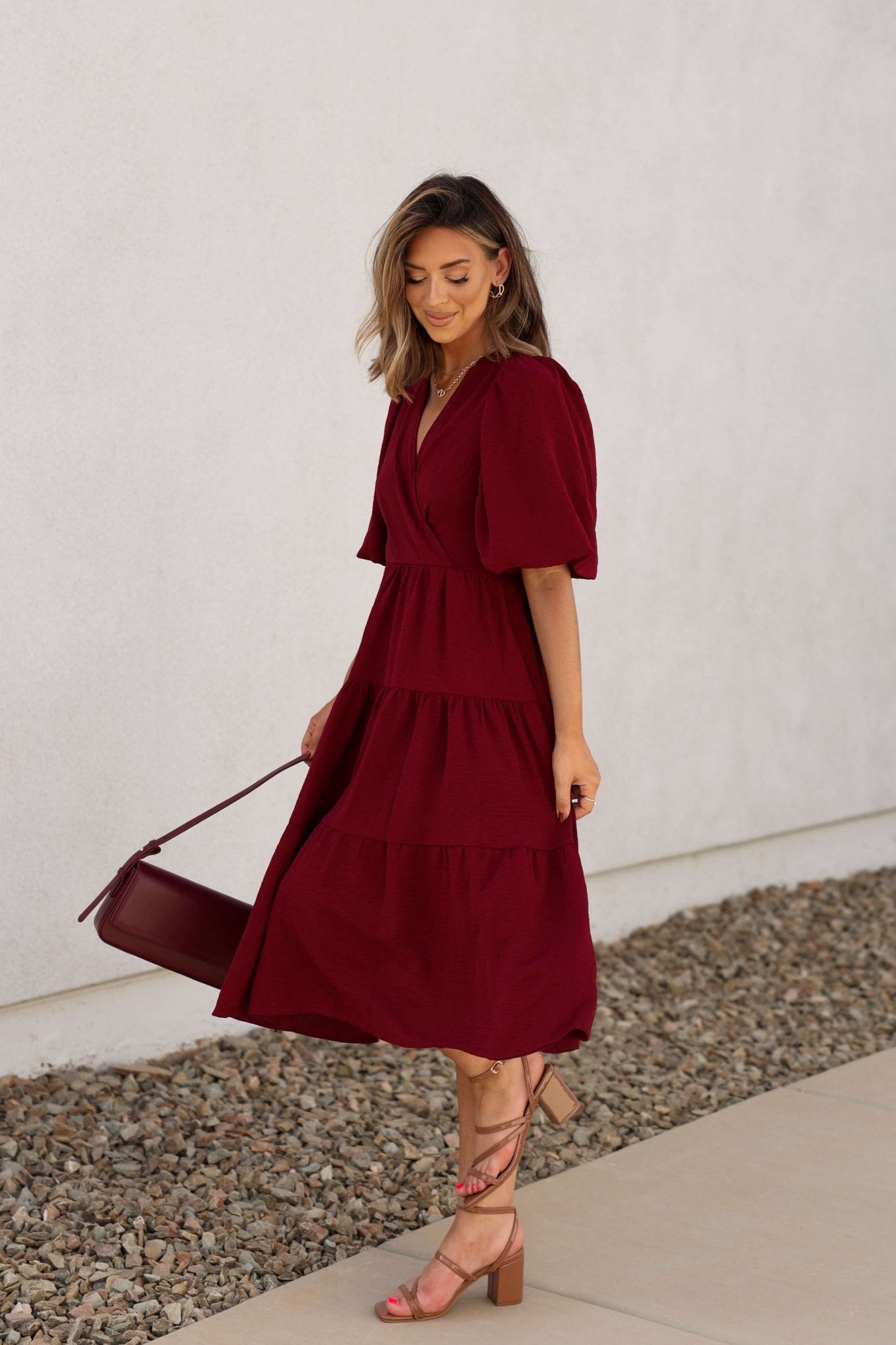 A woman in the Wine Tiered Midi Wrap Dress holds a matching bag and wears strappy heels on a pebble-lined sidewalk.