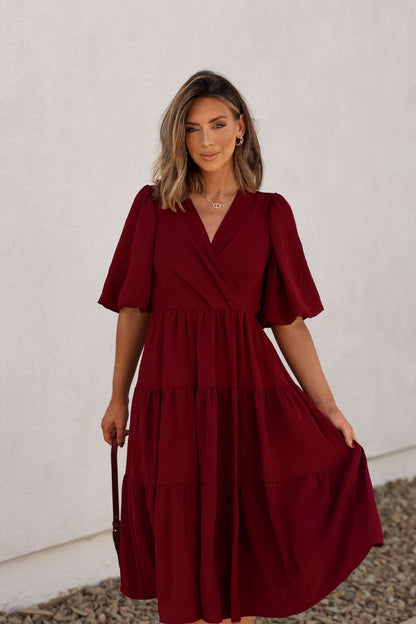Woman wears the Wine Tiered Midi Wrap Dress - FINAL SALE, posing by a white wall and holding the hem of her dress.