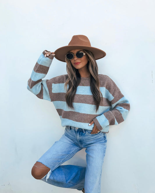 A person wearing an oversized Striped Sweater - Brown And Blue, a fedora, and sunglasses, posed against a white wall.