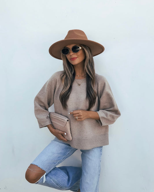A person wears a taupe 3/4 Sleeve Sweater, a wide-brim hat, sunglasses, and distressed light-wash jeans while holding a clutch.
