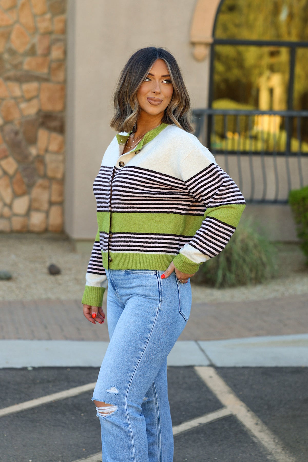 Woman in WISHLIST WL26-13527 Avocado Striped Button Front Cardigan and ripped jeans poses outdoors, smiling sideways.