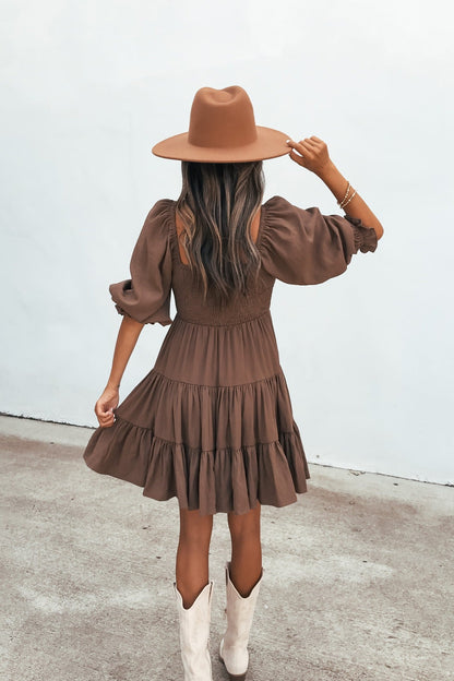 Woman in the Wrenley Tiered Mini Dress - Brown, wide-brim hat, and white boots stands on pavement, back to camera.