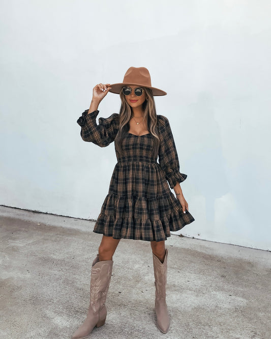 A woman wears a Plaid Mini Dress - Black And Tan with a wide-brimmed hat, sunglasses, and tall boots.