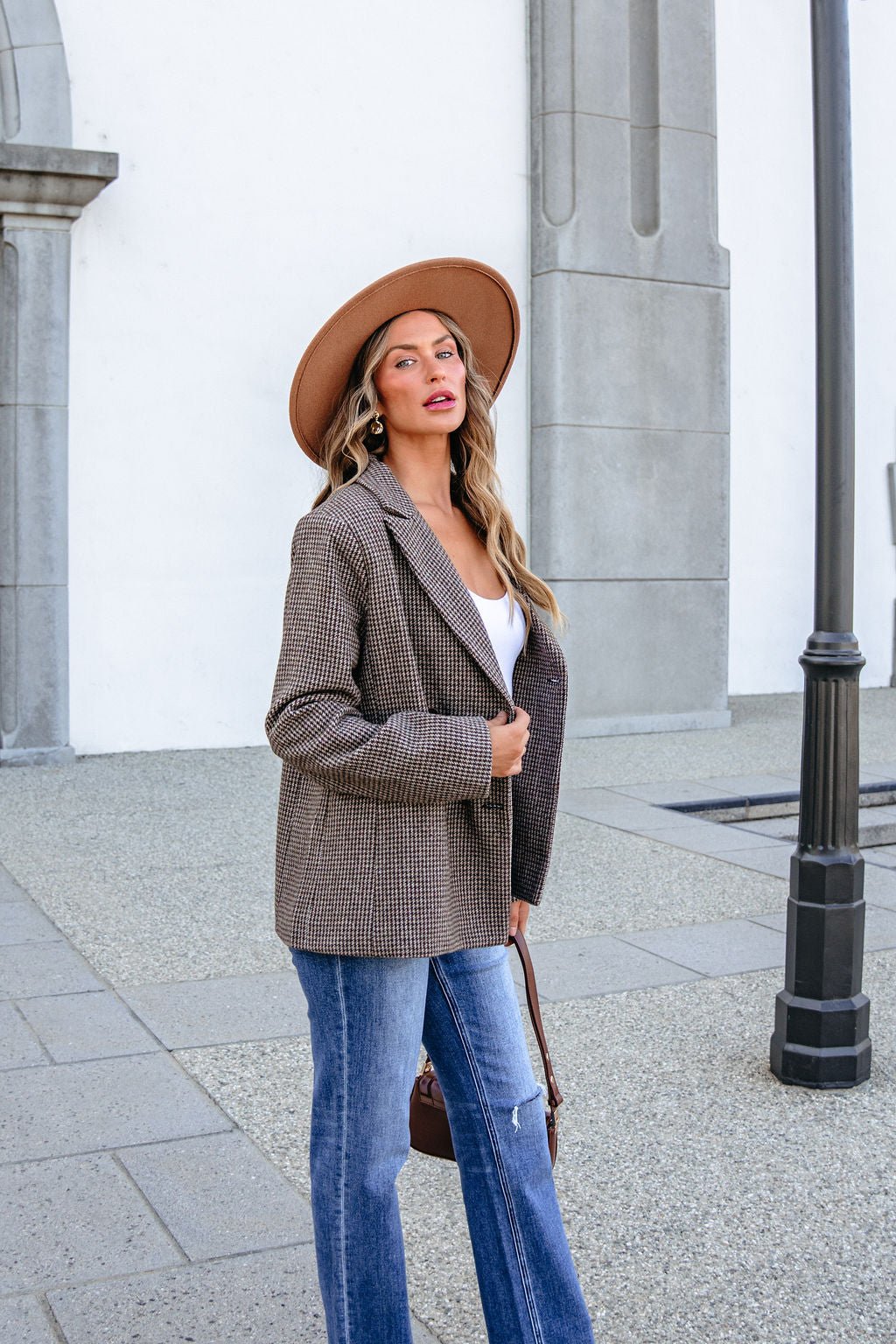 A woman wears a YURA YJ9036BJ BROWN DOUBLE BREASTED HOUNDSTOOTH BLAZER over a white top and blue jeans outdoors.