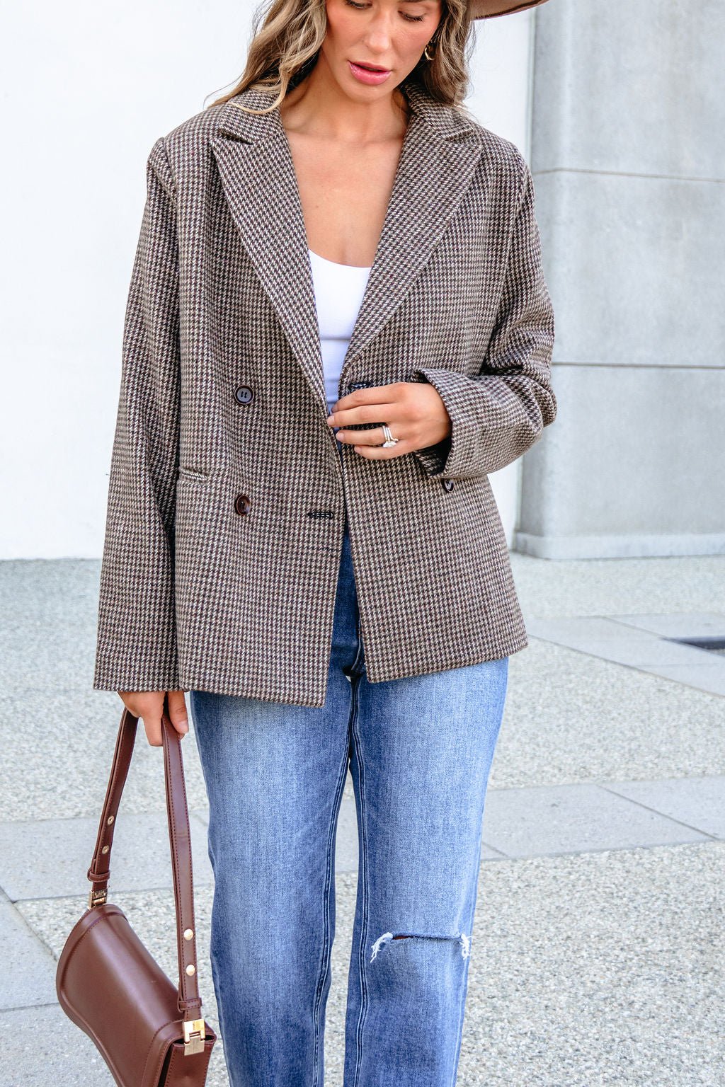 A woman wearing a YURA YJ9036BJ BROWN DOUBLE BREASTED HOUNDSTOOTH BLAZER, white top, and blue jeans, carrying a brown handbag.