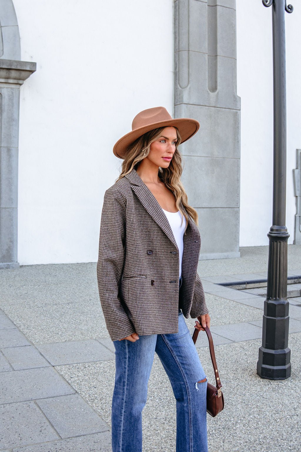 A woman wearing a brown hat and a YURA YJ9036BJ BROWN DOUBLE BREASTED HOUNDSTOOTH BLAZER with blue jeans stands outside.