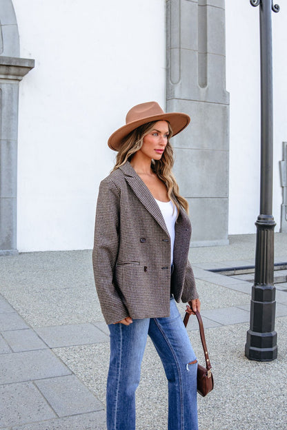 A woman wearing a brown hat and a YURA YJ9036BJ BROWN DOUBLE BREASTED HOUNDSTOOTH BLAZER with blue jeans stands outside.