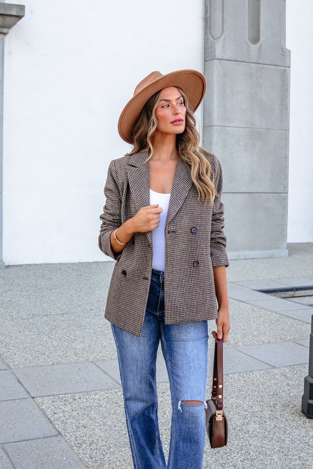 A woman wearing a tan hat, a YURA YJ9036BJ BROWN DOUBLE BREASTED HOUNDSTOOTH BLAZER over a white top, and blue jeans holds a handbag.