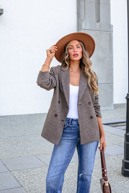 A woman wearing a brown hat, a YURA YJ9036BJ BROWN DOUBLE BREASTED HOUNDSTOOTH BLAZER, a white top, and blue jeans holds a handbag.