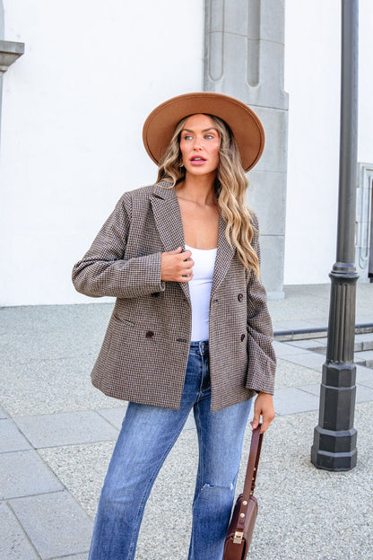 A woman wearing a brown hat, a YURA YJ9036BJ BROWN DOUBLE BREASTED HOUNDSTOOTH BLAZER, and blue jeans holds a handbag.
