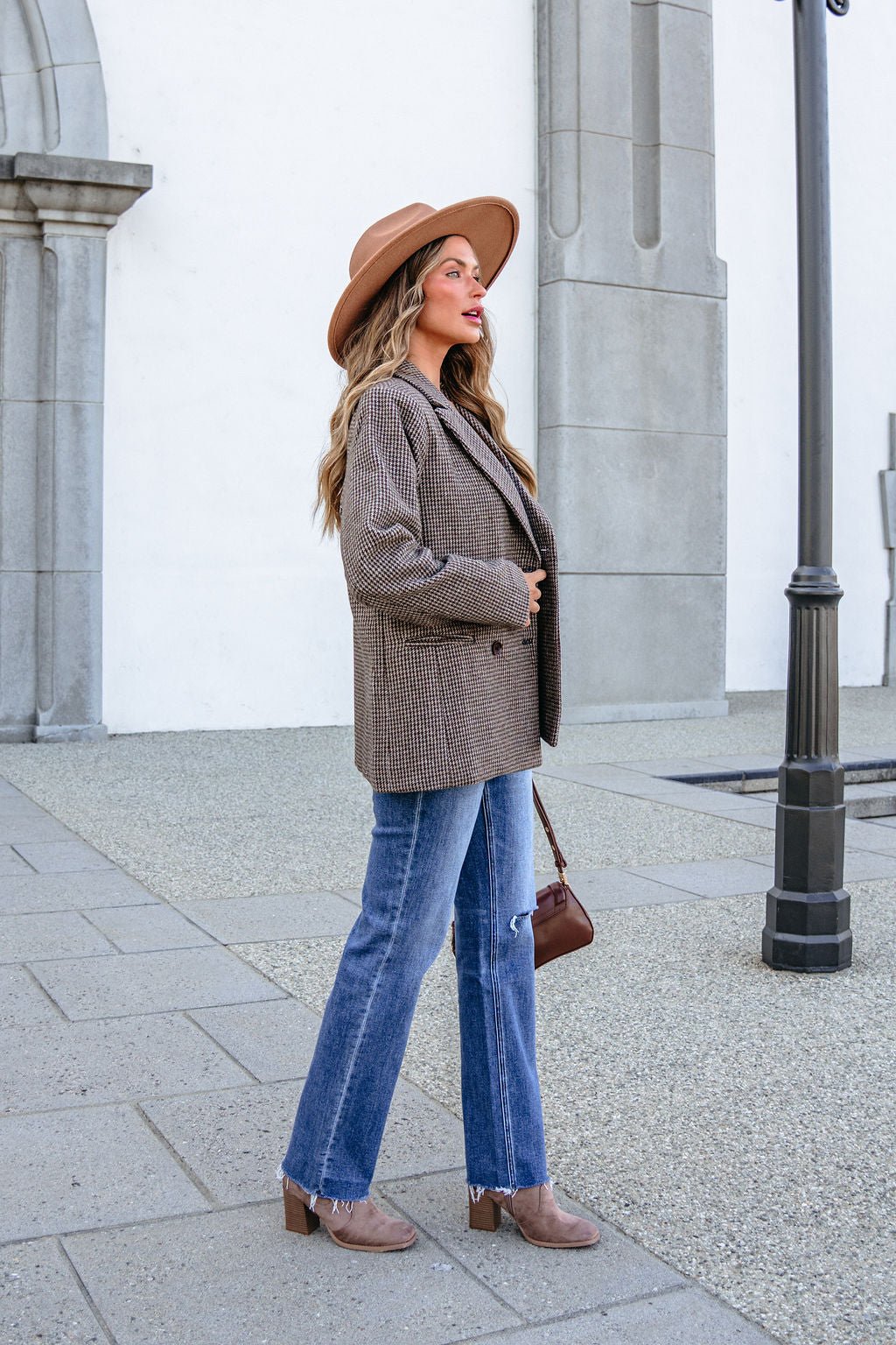 A woman wearing a YURA YJ9036BJ BROWN DOUBLE BREASTED HOUNDSTOOTH BLAZER, brown hat, and blue jeans standing outdoors.