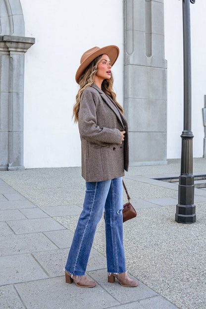 A woman wearing a YURA YJ9036BJ BROWN DOUBLE BREASTED HOUNDSTOOTH BLAZER, brown hat, and blue jeans standing outdoors.