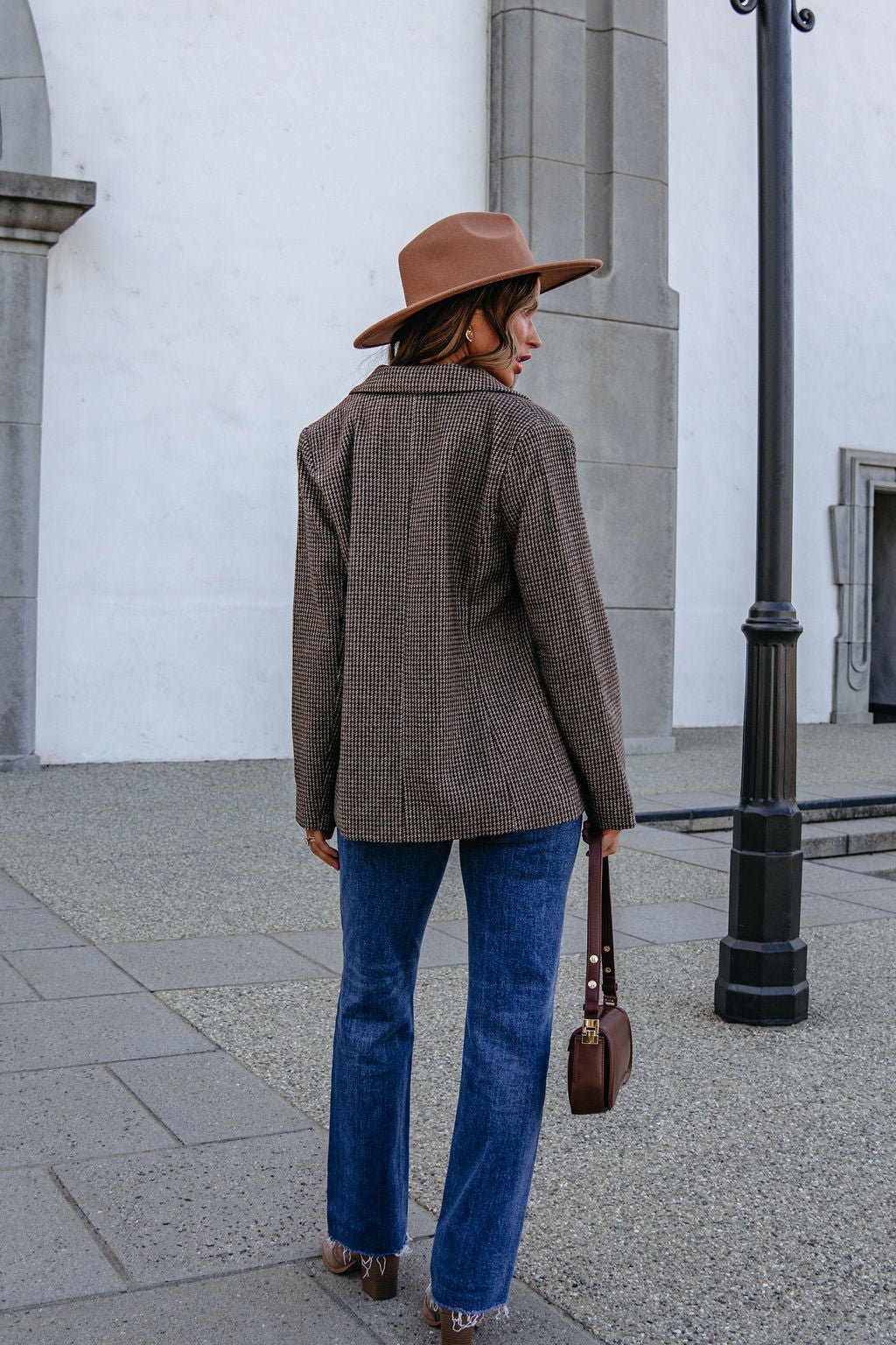 A woman seen from behind, wearing a brown fedora, a YURA YJ9036BJ BROWN DOUBLE BREASTED HOUNDSTOOTH BLAZER, and blue jeans with a shoulder bag.