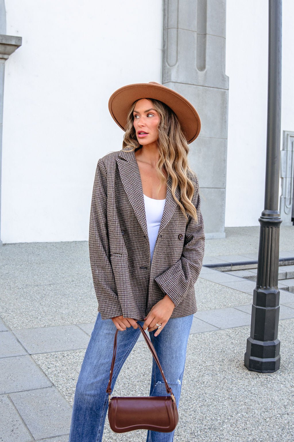 A woman wearing a brown hat, YURA YJ9036BJ BROWN DOUBLE BREASTED HOUNDSTOOTH BLAZER, and blue jeans holds a small brown handbag.