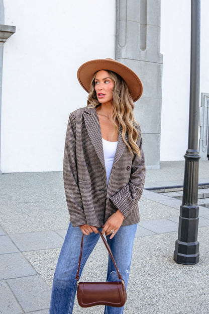 A woman wearing a brown hat, YURA YJ9036BJ BROWN DOUBLE BREASTED HOUNDSTOOTH BLAZER, and blue jeans holds a small brown handbag.