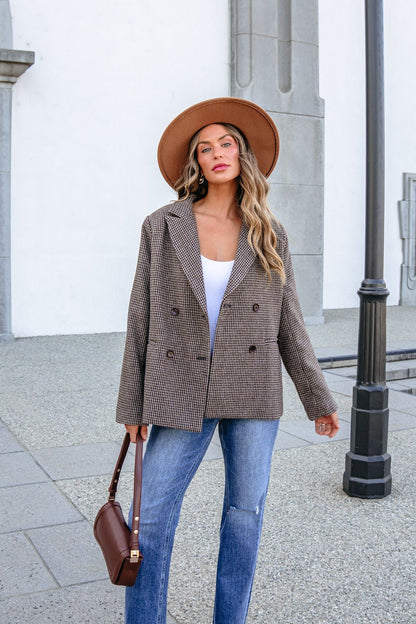A woman wearing a brown hat, YURA YJ9036BJ BROWN DOUBLE BREASTED HOUNDSTOOTH BLAZER, and blue jeans holds a handbag.