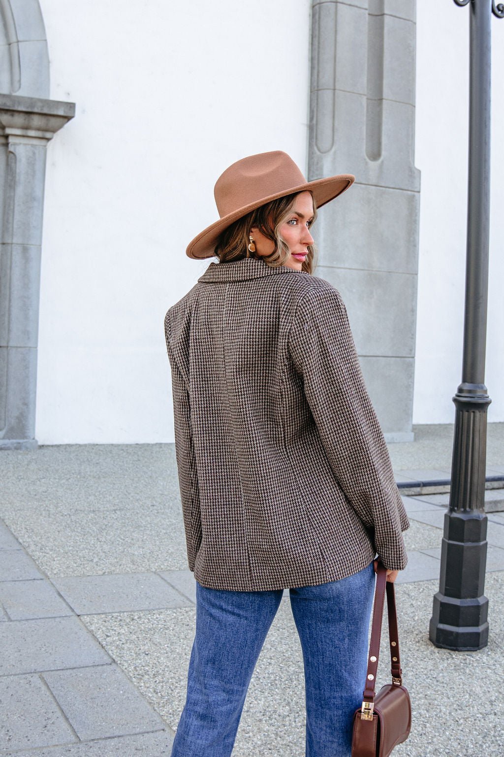 A woman in a brown hat and YURA YJ9036BJ BROWN DOUBLE BREASTED HOUNDSTOOTH BLAZER looks over her shoulder while carrying a handbag.