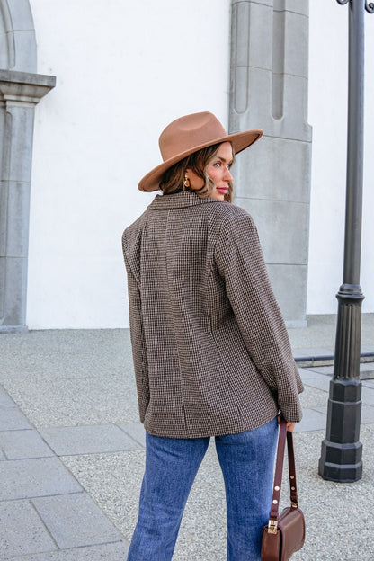 A woman in a brown hat and YURA YJ9036BJ BROWN DOUBLE BREASTED HOUNDSTOOTH BLAZER looks over her shoulder while carrying a handbag.